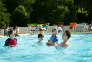 Family with children playing in the busy pool at the Hersheypark Camping Resort. Other guests laying on lounge chairs by the pool.