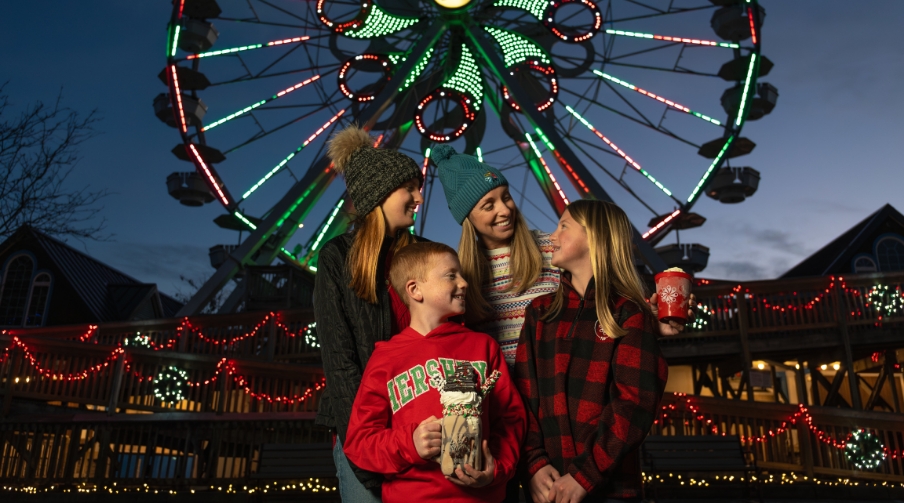 Family standing in front of the Ferris Wheel at Hersheypark lit up red and green.