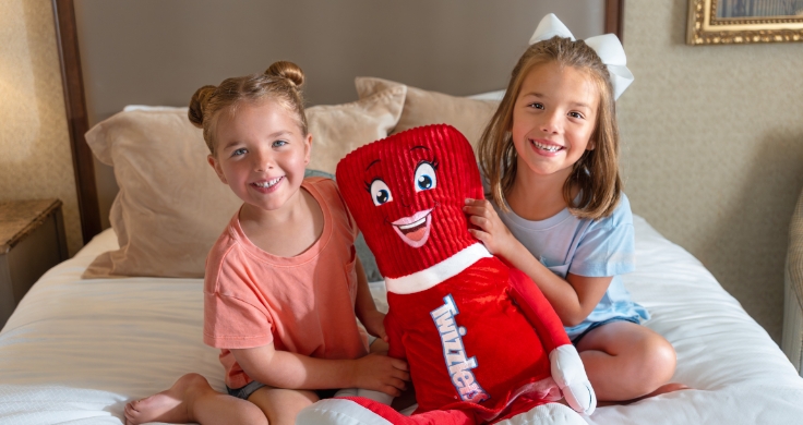 Two young sisters sitting on a bed at the Hershey Lodge and holding up their stuffed Twizzler.